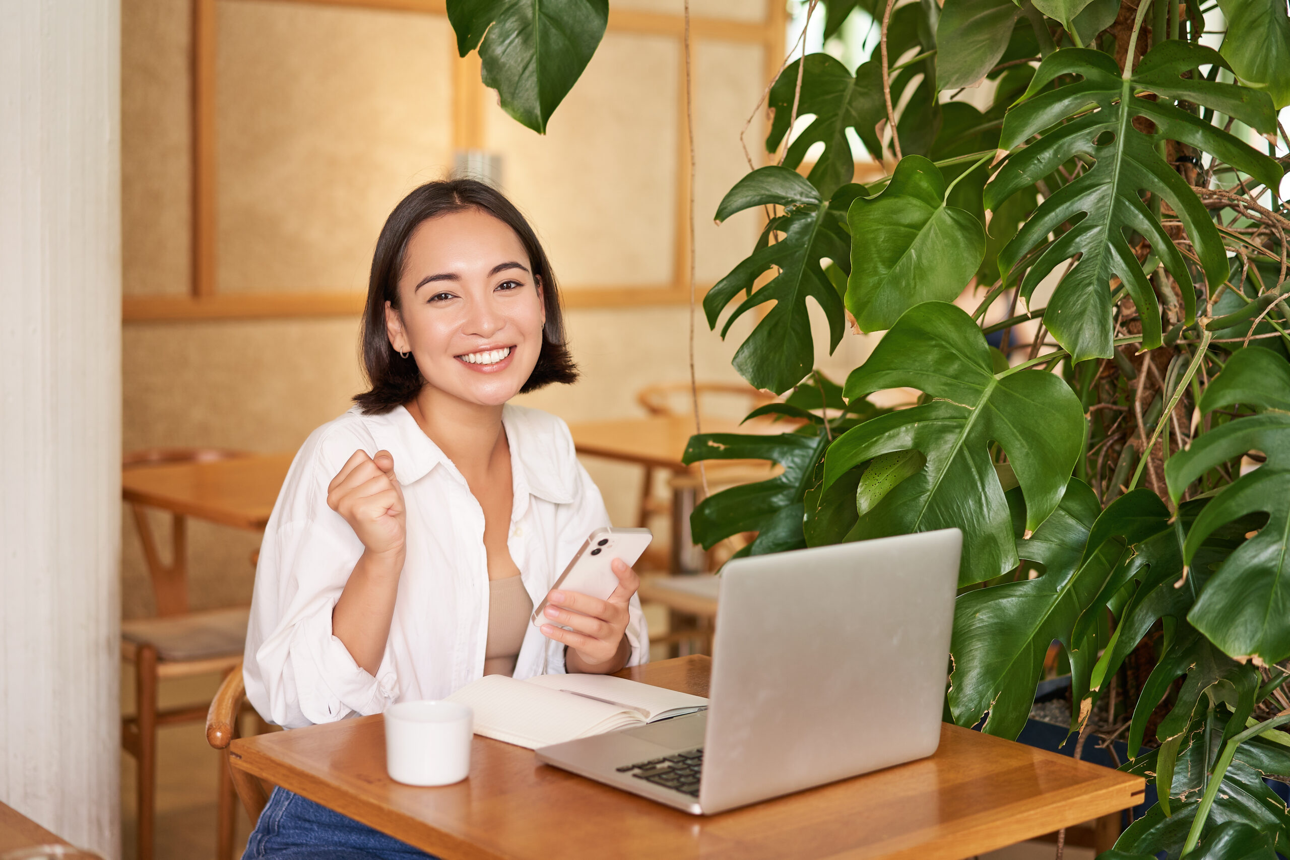 Happy young asian woman feel success, winning and celebrating, sitting with laptop and smartphone in cafe and triumphing, shouting yes